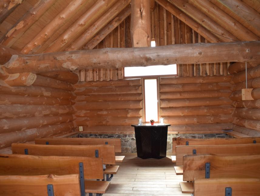 Blick ins Innere der Kapelle mit Holzbänken, Altar, einem dicken Querbalken, der das Dach mit trägt und dem Fenster in Form eines Kreuzes