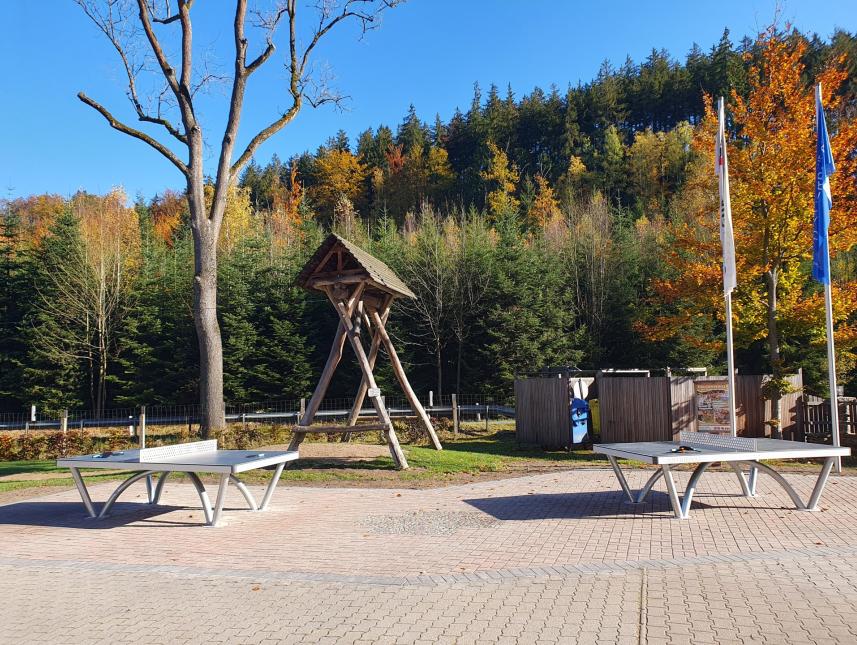 zwei Tischtennisplatten warten auf dem Freigelände auf Spieler; die Gastfreundefahne und die Fahne mit dem Logo der himmlischen Herbergen hängen mangels Wind schlaff am Mast