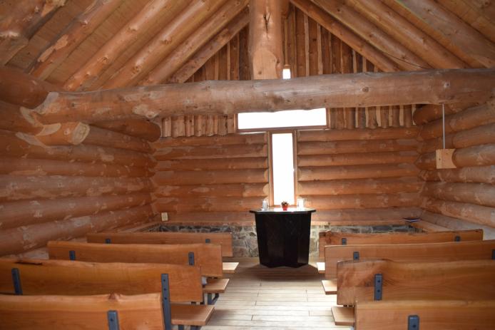 Blick ins Innere der Kapelle mit Holzbänken, Altar, einem dicken Querbalken, der das Dach mit trägt und dem Fenster in Form eines Kreuzes