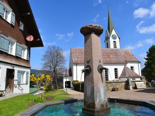 Dorfbrunnen, dahinter die Kirche St. Anna 